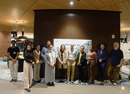 Group of a dozen people standing in front of wall of wood paneling.